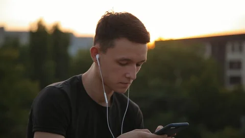 Handsome young man in earphones using phone at sunset outdoors 2 Video stock 94621873