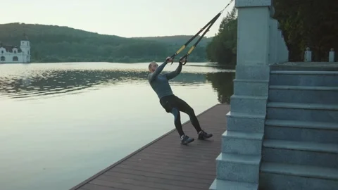 Handsome young man exercising with trx suspension strap near lake crossfit Stock Footage 138650455