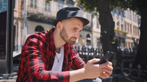 Handsome young man in hat uses phone sitting in city look at camera smile happy Stock Footage 92071936