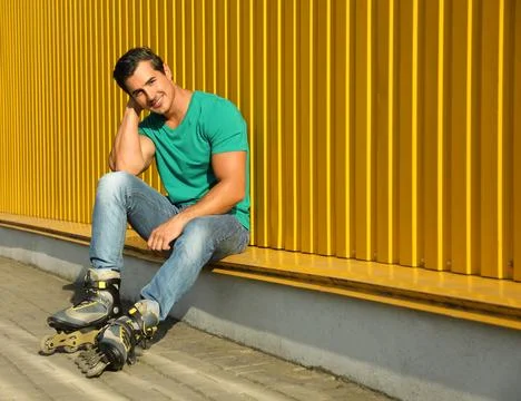 Handsome young man with inline roller skates sitting near yellow building, sp Stock Photos