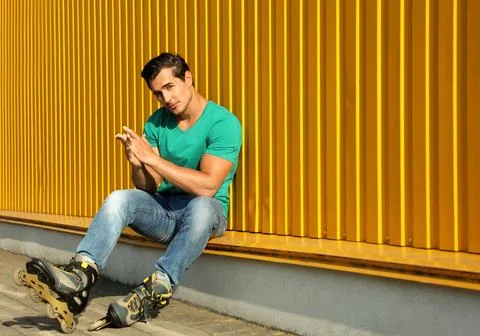 Handsome young man with inline roller skates sitting near yellow building, sp Fotos de archivo