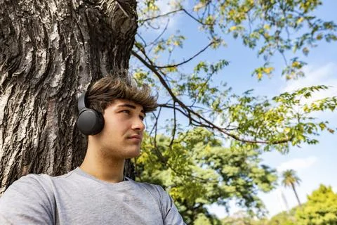 A handsome young man leaning on a tree and listening to music. It seems he .. Stock Photos