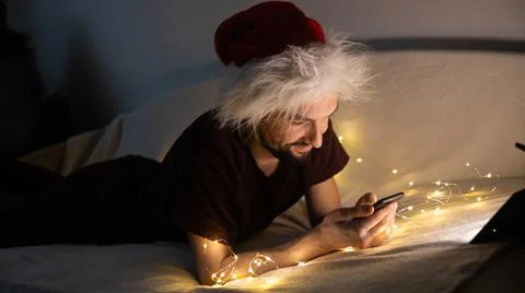Handsome young man lying on the couch wearing santa hat, using his phone and Stock Photos