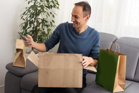 Handsome young man opening box with parcel while sitting on sofa at home. Stock Photos
