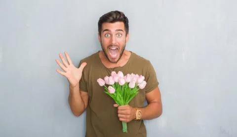Handsome young man over grey grunge wall holding flowers bouquet very happy a Stock Photos