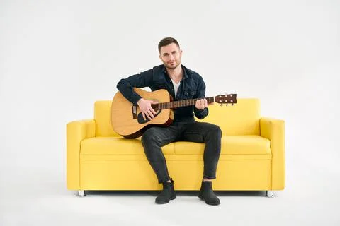 Handsome young man playing acoustic guitar whlile sitting on yellow sofa over Stock Photos