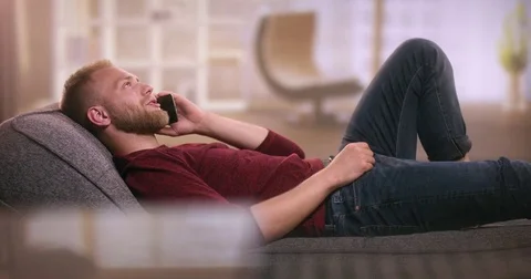 Handsome young man relaxing on sofa and talking on smartphone Stock Footage 77413778
