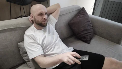 Handsome young man with remote control in his hands sitting on couch at home Stockbeeldmateriaal 311152288