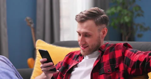 A handsome young man sits on couch in a room and uses the phone. 20s man Stockbeeldmateriaal 153897763