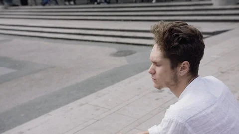 A handsome young man sitting down and looking concerned in a city square. Stock Footage 115981402