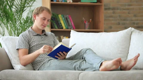 Handsome young man sitting on sofa reading book looking at camera and smiling Stock Footage 34278339