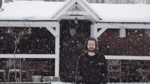 Handsome young man standing by an old wooden house during snowfall. 動画素材 60736934