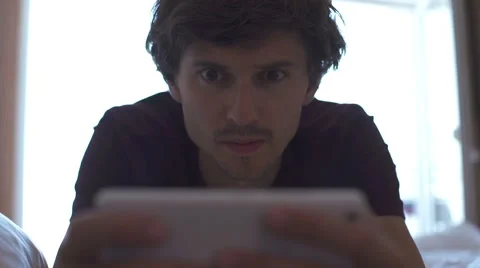 Handsome young man with stubble, relaxing in bed at home and reading an ebook on Stock Footage
