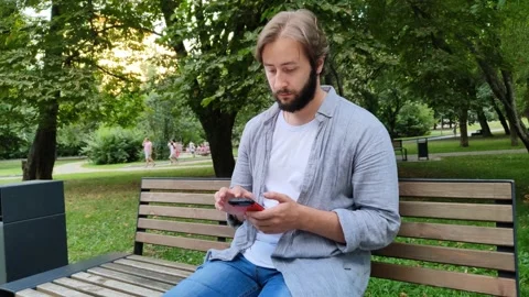 Handsome young man use mobile phone while sitting on bench in public park Video stock 215079634