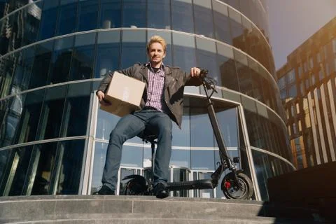 A handsome young man uses an electronic scooter to deliver goods in a cardboa Stock Photos