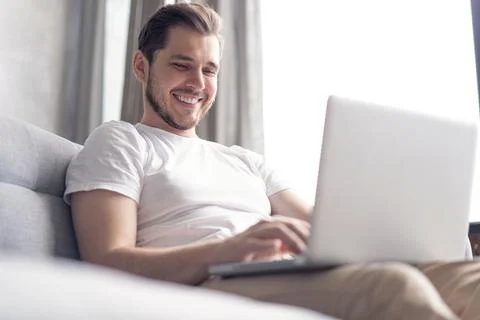Handsome young man using laptop with smile while sitting on the couch at home Stock Photos