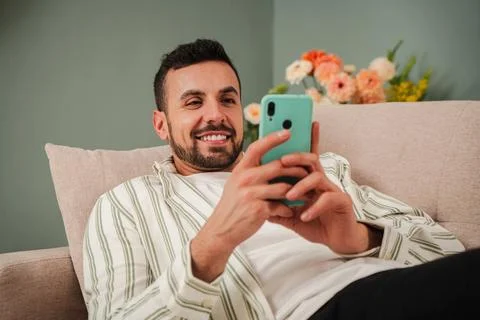 Handsome young man using a mobile phone lying on a sofa at his living room Stock Photos