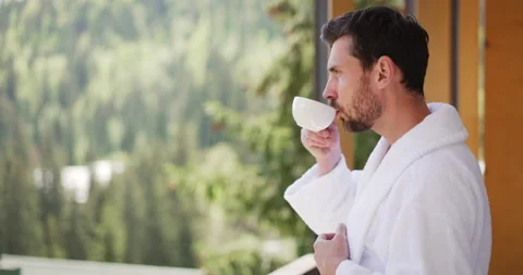Handsome young man in white bathrobe standing on balcony of hotel room, drinking Stock-Footage 156969091