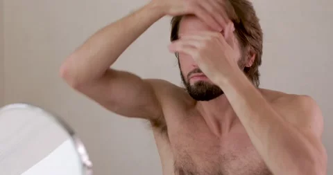 Handsome young man without shirt doing styling for hair and beard while looking Stock Footage 142528362