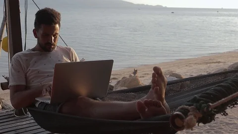 A handsome young man working on his laptop in the hammock on the beach. Stock Footage 99891696