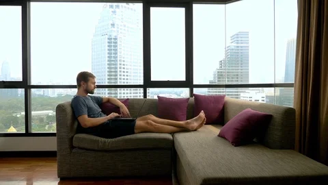 Handsome young man working on a laptop on a couch in front of a panoramic window Stock Footage 101866057