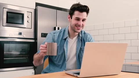 Handsome young man working with a laptop and smiling at the kitchen with a cup Stock Footage 248609448