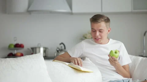 Handsome young men sitting on sofa reading book and eating apple Stock Footage 27692060