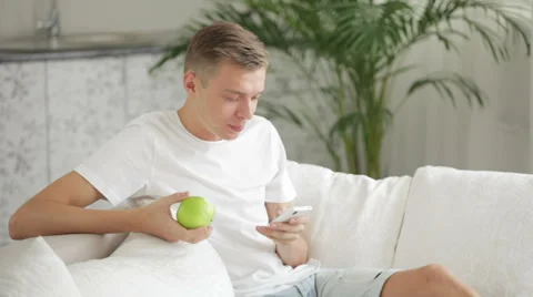 Handsome young men sitting on sofa eating apple and using cellphone Stock Footage 27692071