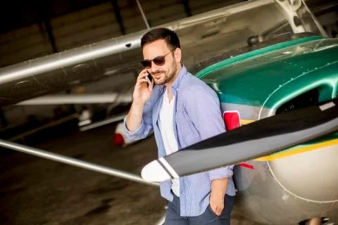 Handsome young pilot checking airplane in the hangar and using mobile phone Stock Photos