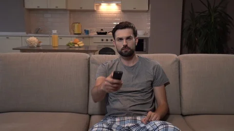 Hansdsome man sits on sofa and watches TV. He uses remote control several times Stock Footage 99739156