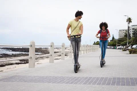 Happy afro african american young couple riding push scooters by beach in city, Stock Photos