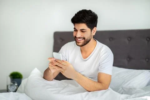 Happy arab guy using smartphone in bed, waking up in the morning and browsing Stock Photos