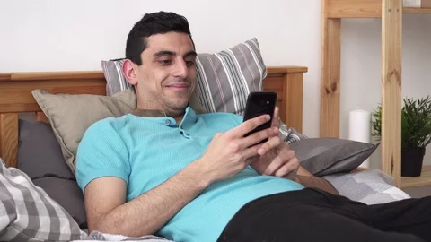 Happy arabic man using a smartphone, lying on bed at home Stock Footage 86402312