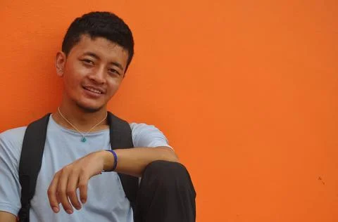 A happy Asian guy looking at camera, sitting against orange wall background 写真素材
