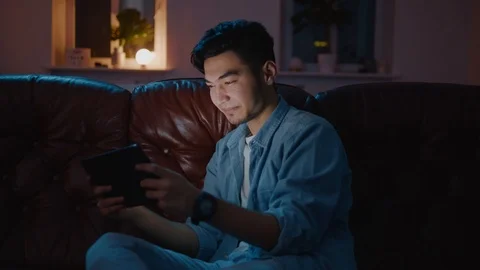 Happy Asian guy scrolling webpages on tablet at night sitting on sofa. Stockbeeldmateriaal 105584439