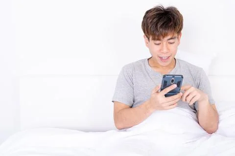 Happy Asian man using a mobile phone lying on the bed at home. Social and tec Stock Photos
