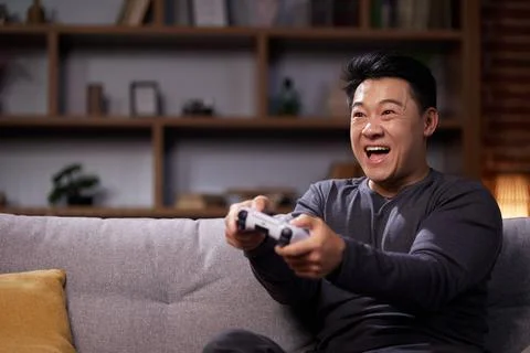 Happy Asian man using a wireless joystick and playing video games at home Stock Photos