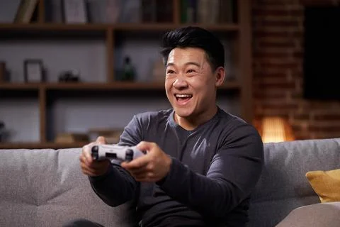 Happy Asian man using a wireless joystick and playing video games at home Stock Photos
