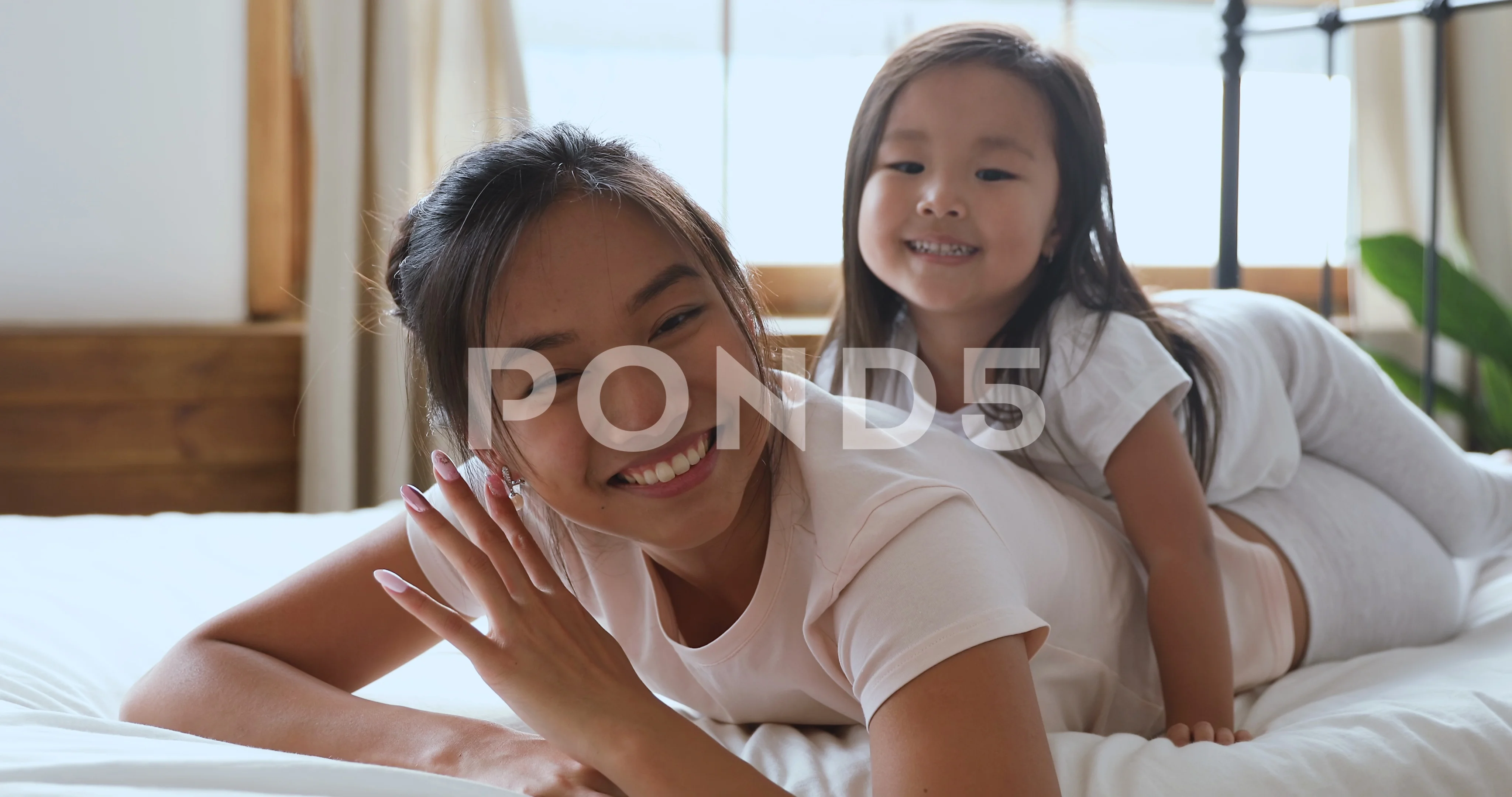 Happy asian mom and daughter looking at camera in bed