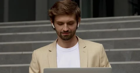 Happy bearded guy using wireless laptop, sitting on stairs, smiling and looking Video stock 137744092