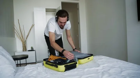 Happy bearded man getting ready for holiday putting in clothes in suitcase at Stock Footage 123528118
