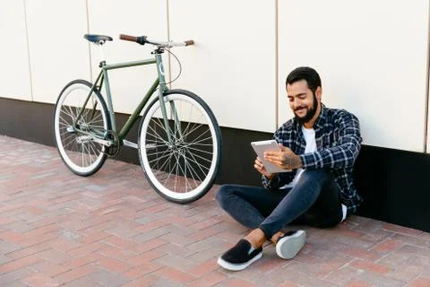 Happy bearded man using a tablet while sitting on the ground near the bike. Stock Photos