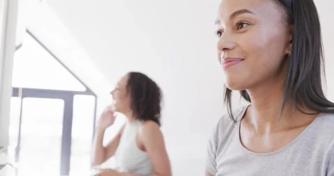 Happy biracial lesbian couple using face cream and talking at mirrors in sunny Video stock 242065630