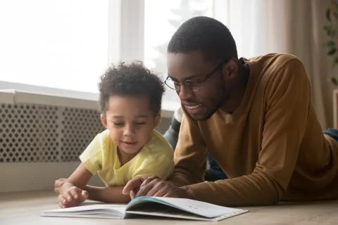 Happy black father and toddler son reading book at home Stock Photos