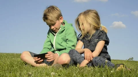 Happy blond children using smartphone sitting on the grass. Brother and sister. 動画素材 63039109