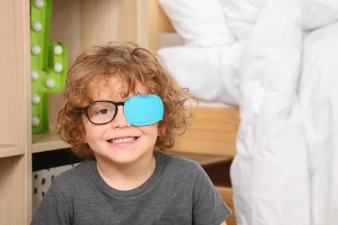Happy boy with eye patch on glasses near shelf indoors. Strabismus treatment Fotos de archivo