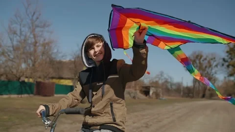 A happy boy with a flying rainbow kite rides a bicycle on the road. Video stock 192300870