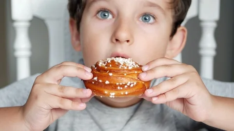 Happy boy is sitting behind the table and eating a cinnamon bun, closeup Stock Footage 97531555