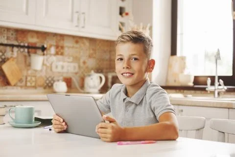 Happy boy sitting with tablet at the table in the home kitchen, chatting on Stock Photos
