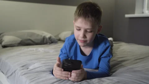 Happy boy talking on a video link on a smartphone, lying at home on a bed Video stock 258339150
